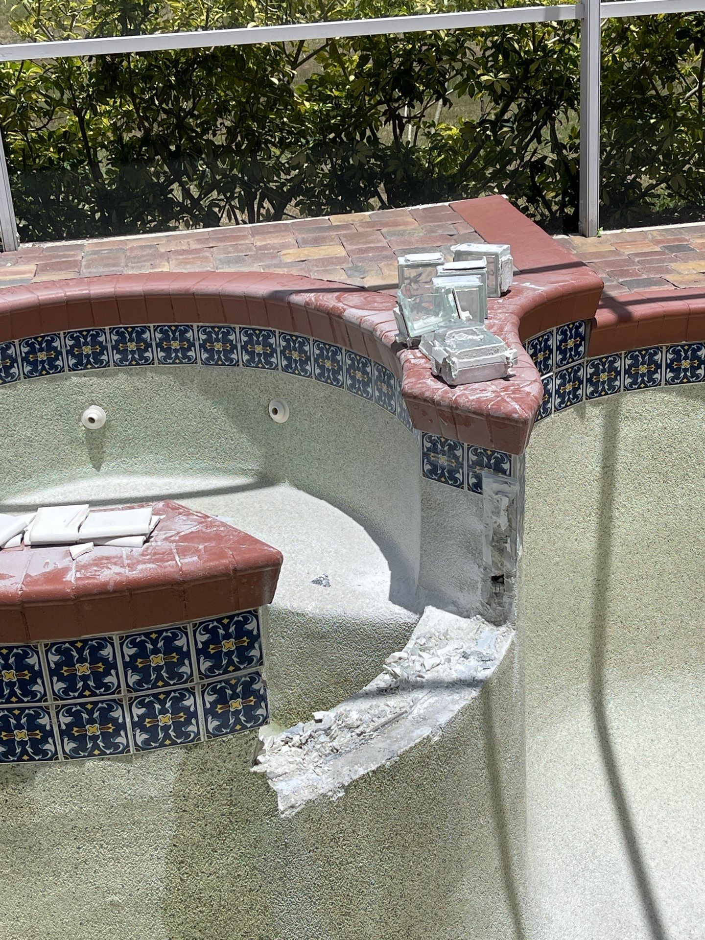 Damaged mosaic pool wall with broken concrete and a metal fixture on the coping.