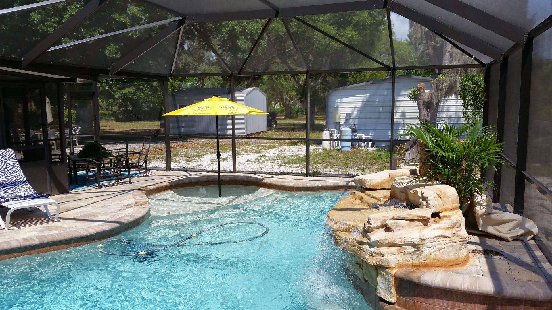 Screened-in backyard pool with blue water, stone waterfall, patio chairs, and a yellow umbrella