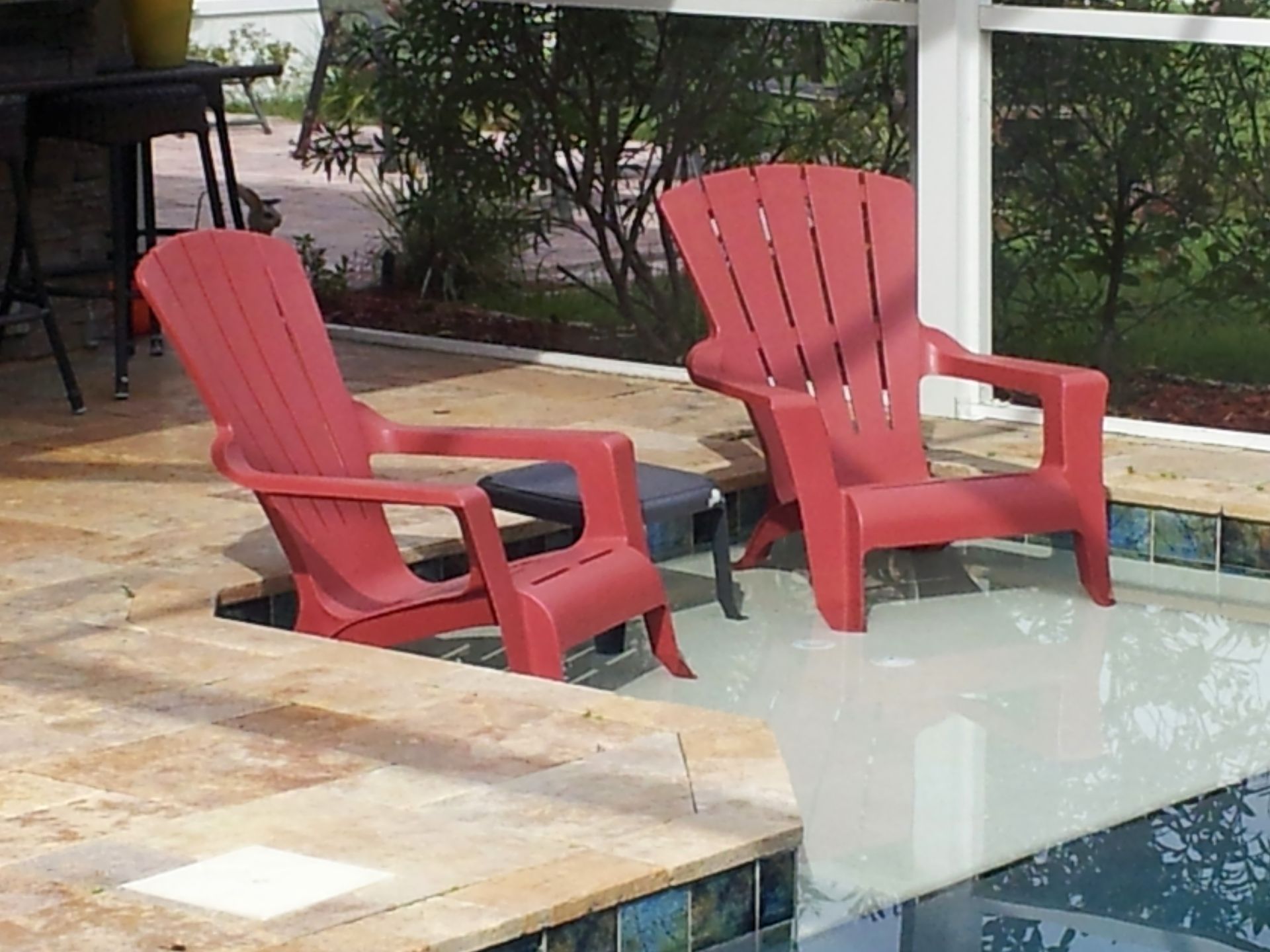 Two red Adirondack chairs beside a pool with a glass tabletop in a patio setting