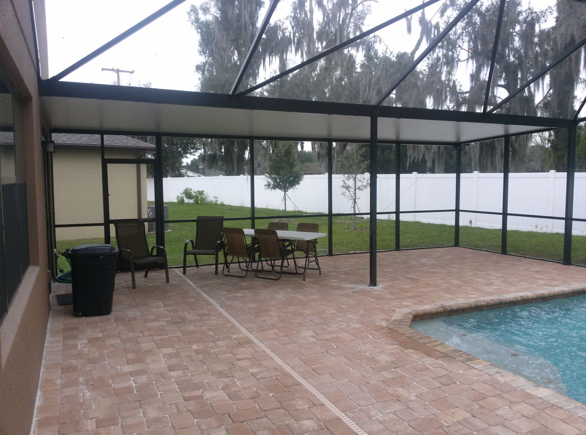 Covered patio with green chairs and table beside a pool, overlooking a lake through screened enclosure