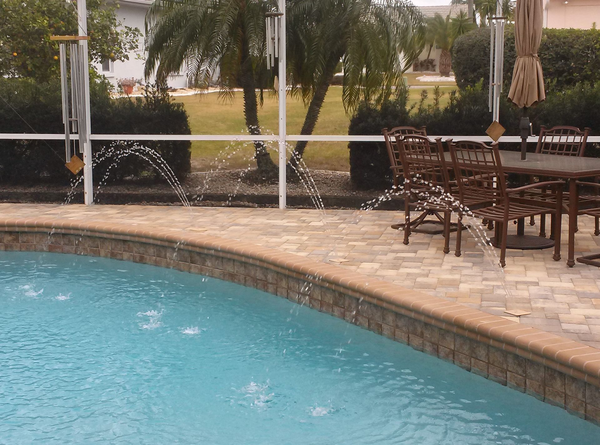 Poolside patio with turquoise water, stone deck, and outdoor tables and chairs under trees