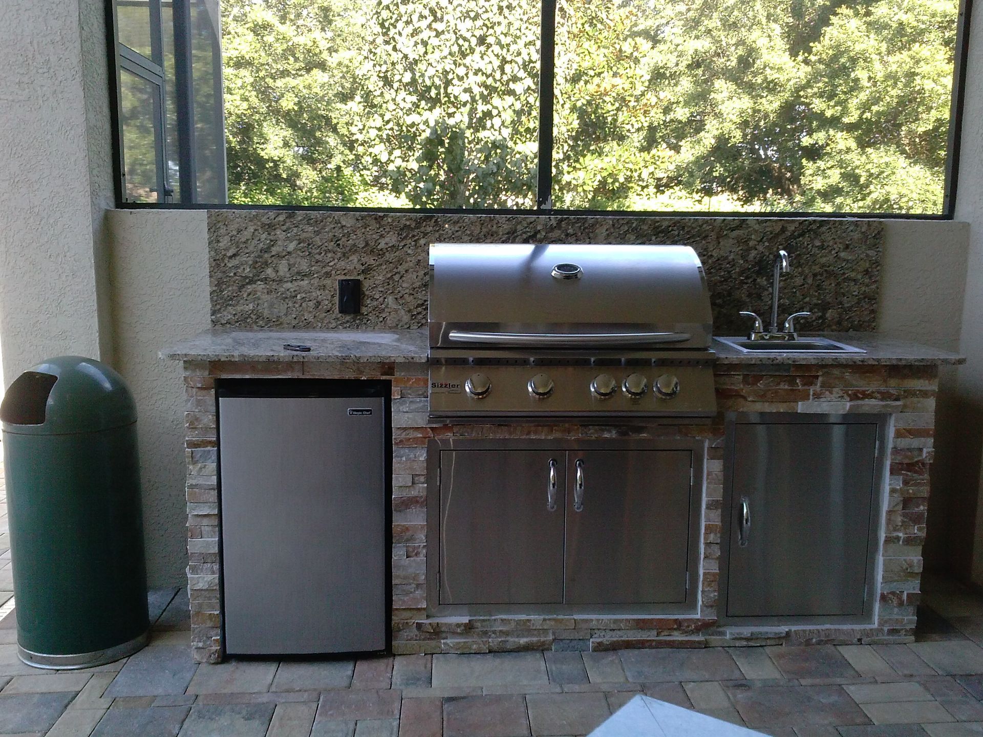 Outdoor kitchen with stainless grill, mini fridge, sink, and green propane tank beneath a large window
