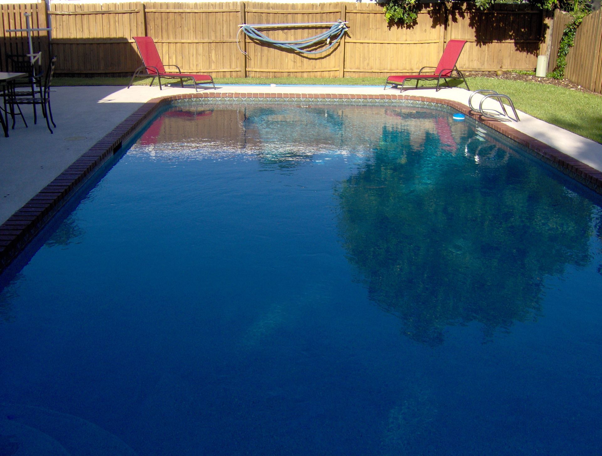 Backyard swimming pool with blue water, patio chairs, and a hammock on a wooden fence