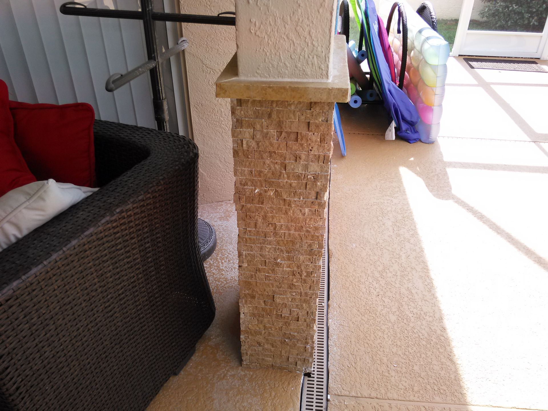 Sunlit patio with wicker chair, stone column, and tiled floor beside a walkway