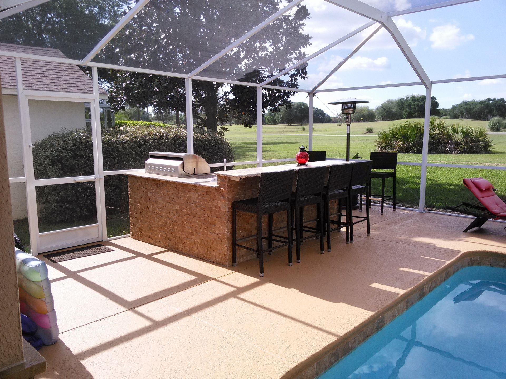 Poolside patio with outdoor bar, high stools, lounge chair, and screened enclosure overlooking a lawn