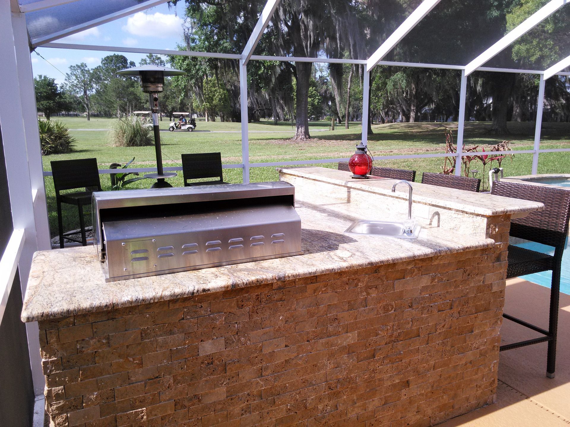 Outdoor kitchen with granite counter, built-in grill, and bar seating under a screened patio overlooking a grassy yard