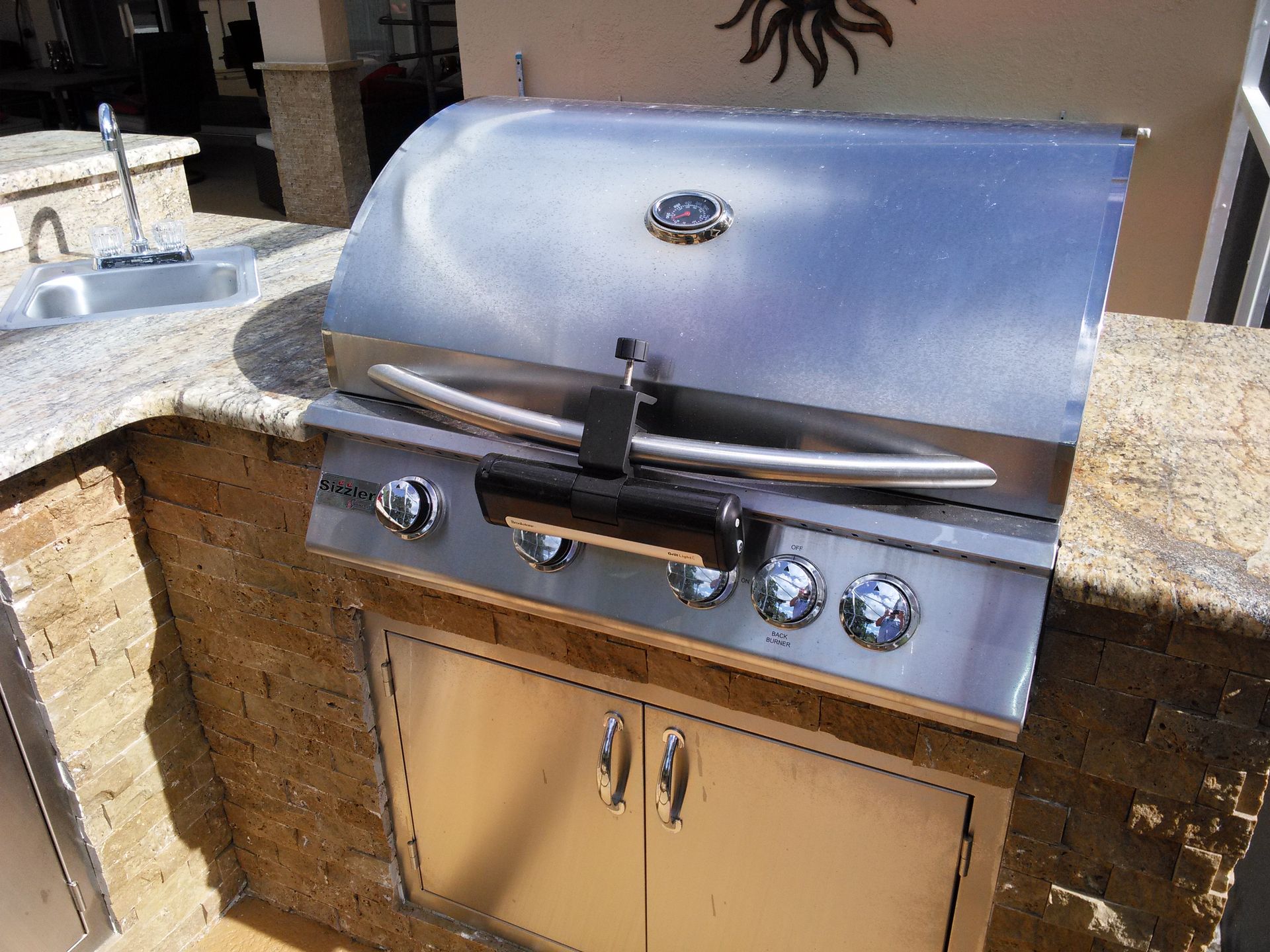 Stainless steel gas grill built into an outdoor stone counter with tan cabinet doors.