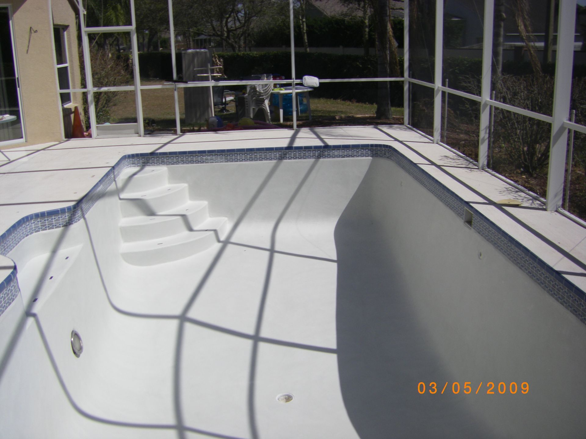 Empty white backyard swimming pool with steps, enclosed by a screen fence under bright sunlight