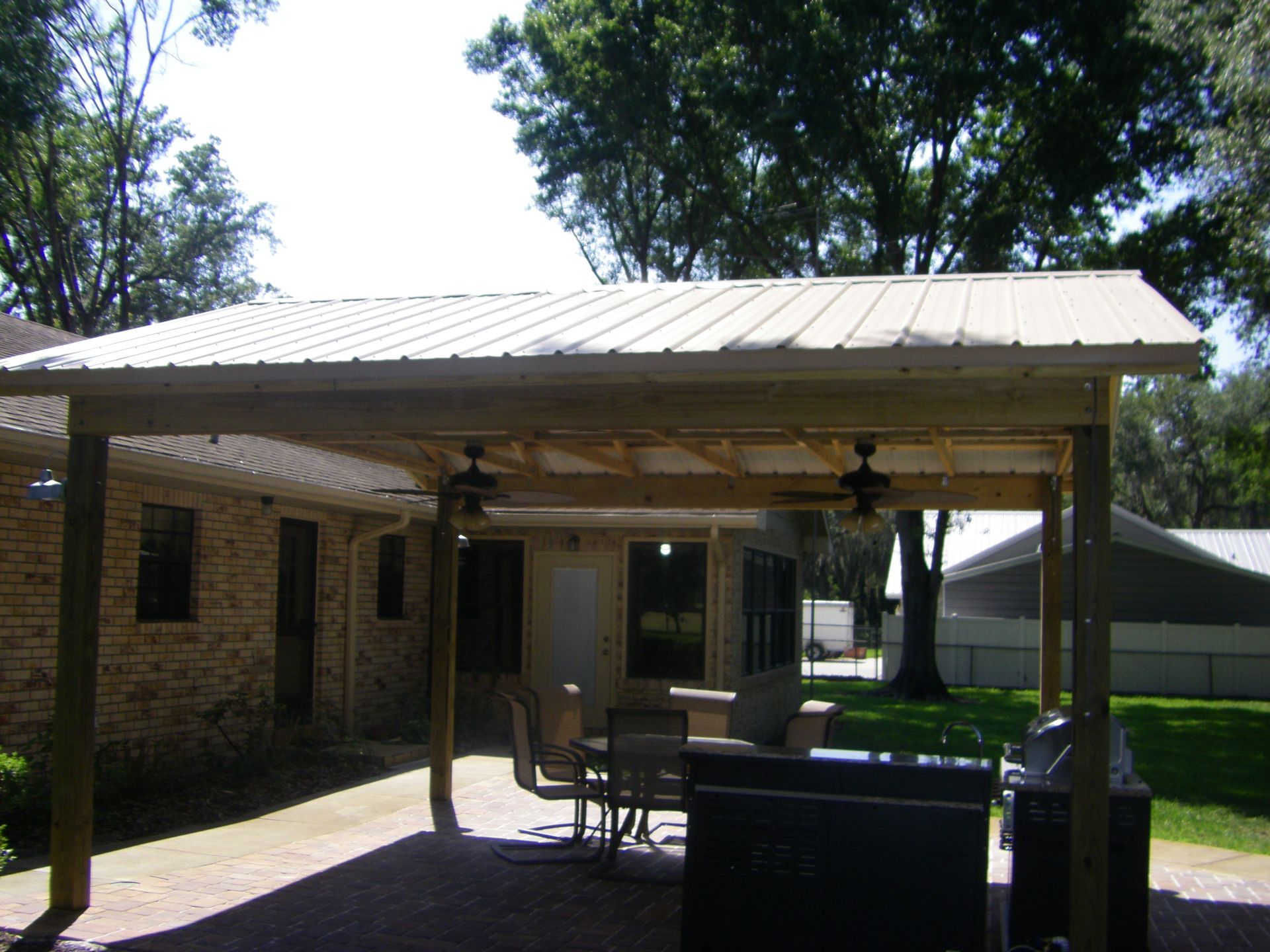 Covered patio with brick house, metal roof, outdoor chairs, and a barbecue grill in a sunny yard