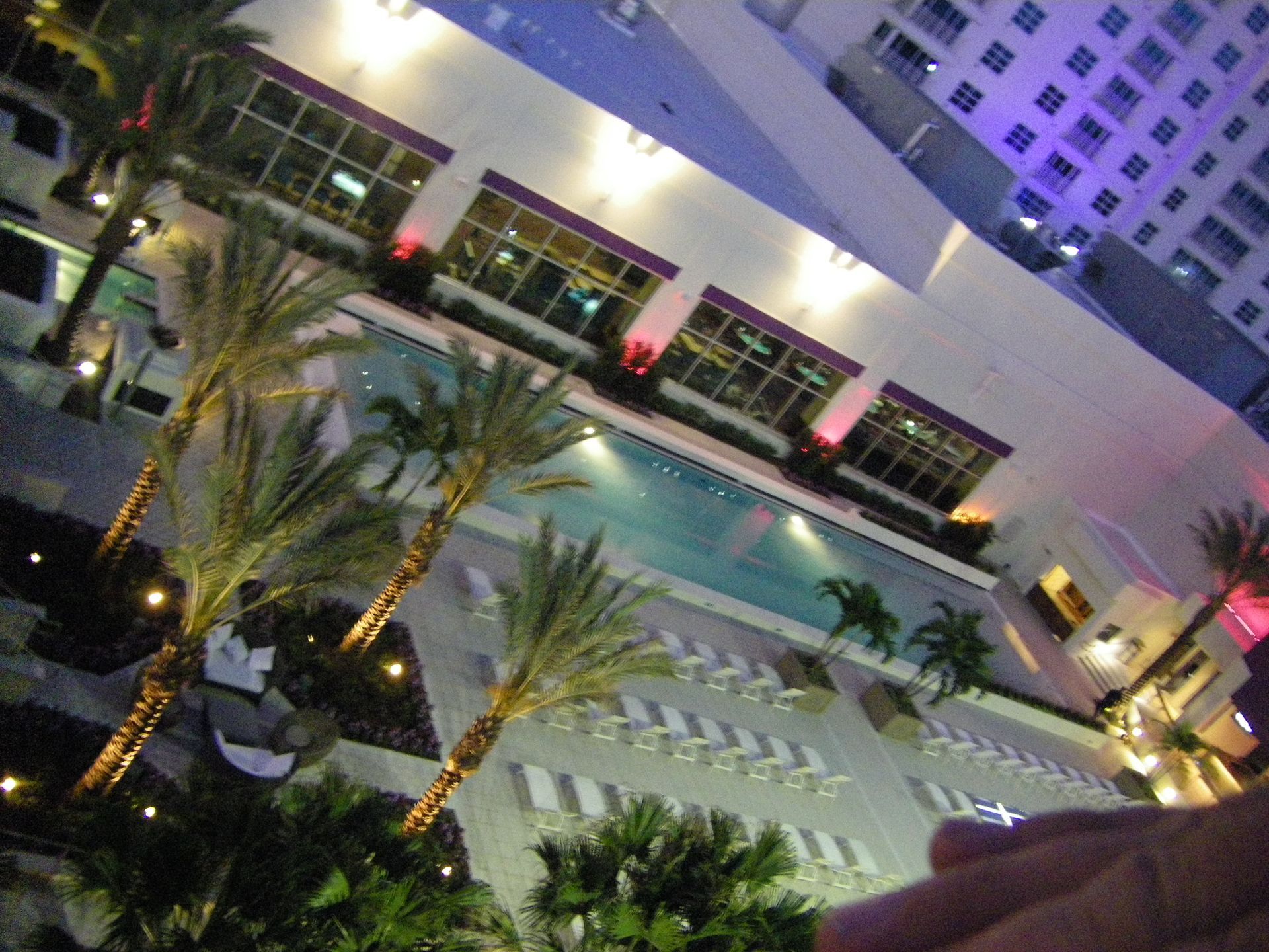 Nighttime view of a lit hotel courtyard with palm trees, pool, and white lounge chairs