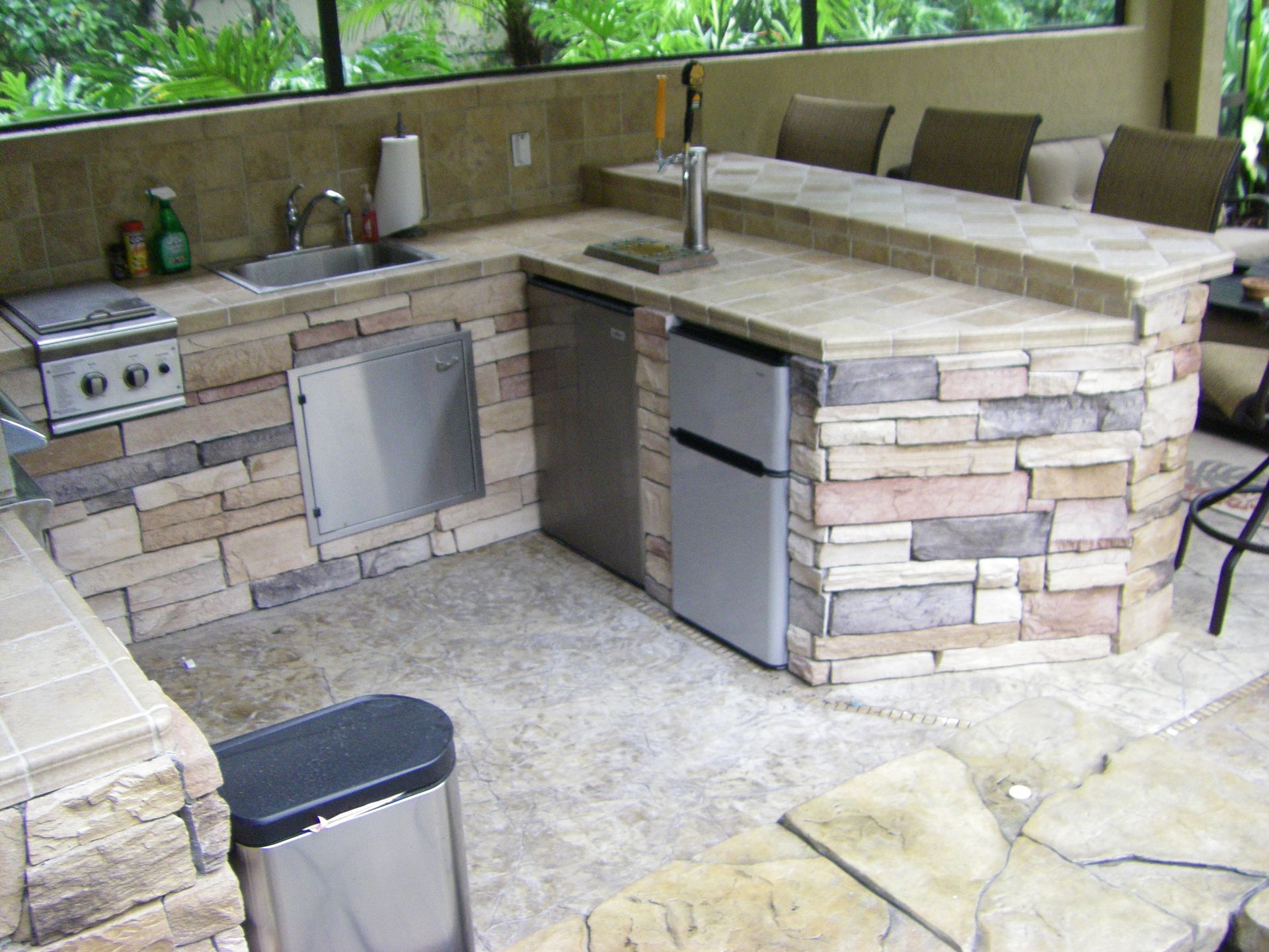 Outdoor stone patio kitchen with sink, grill, cabinets, and stone counters under a covered porch