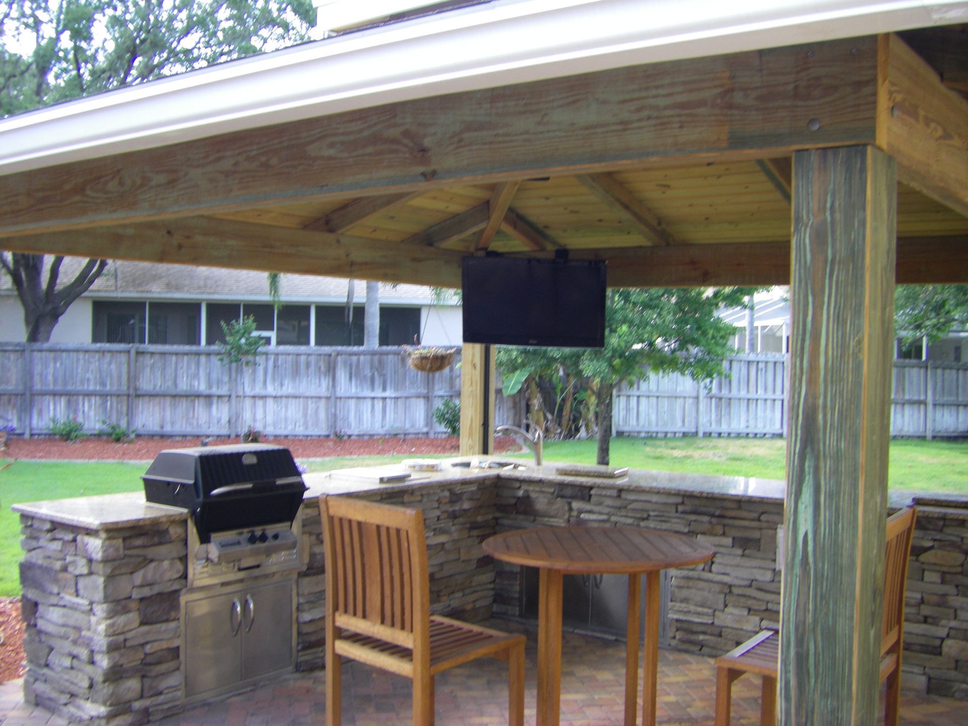 Covered backyard patio with stone grill, wooden table and chairs, and a fenced lawn beyond.