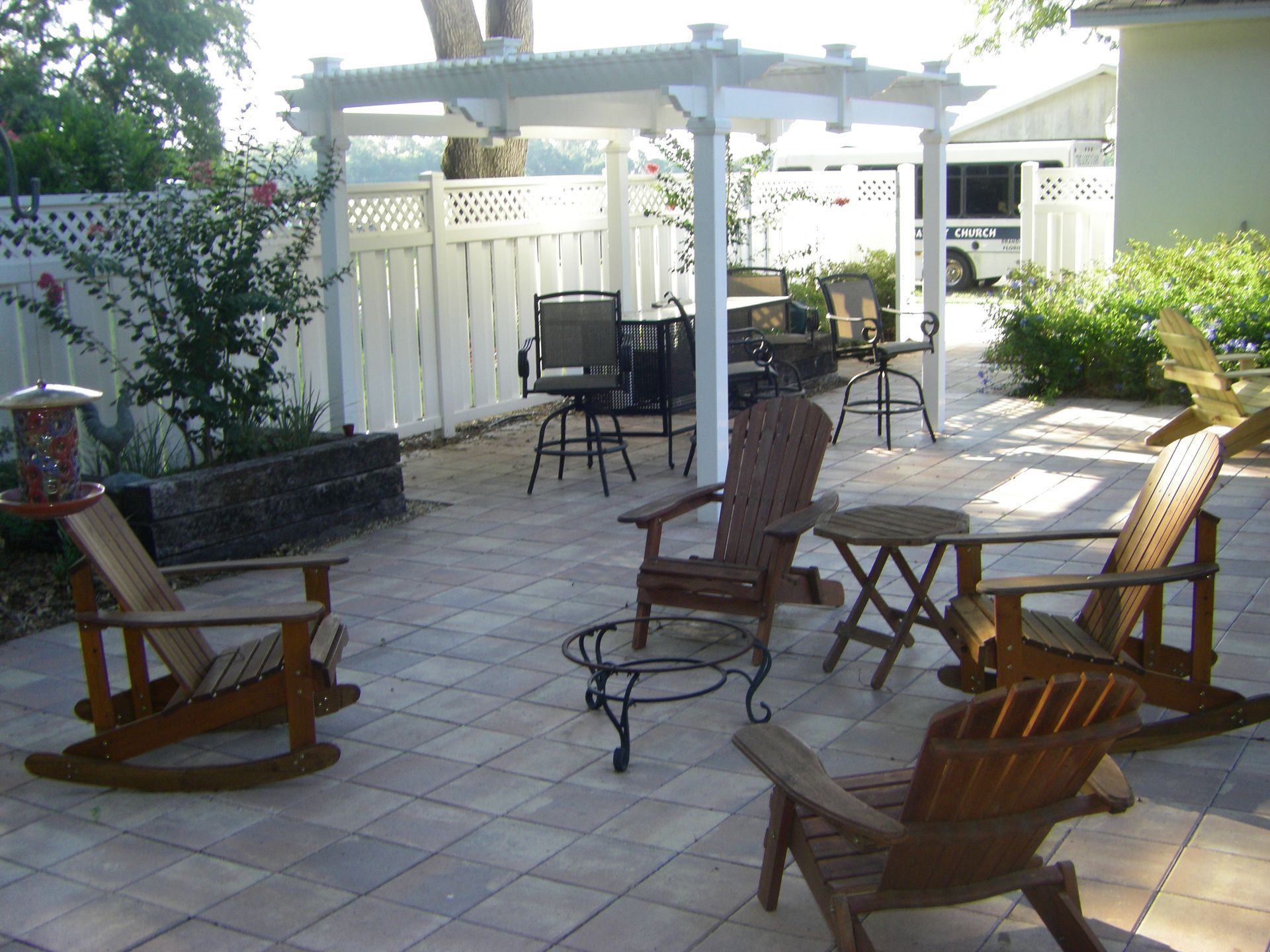 Patio with wooden chairs around a paved courtyard, white pergola, and potted plants in sunlight