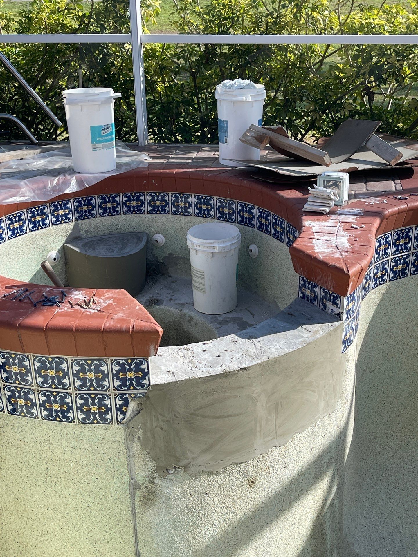 Outdoor tiled fountain under construction with buckets and tools on top, brick edging, and white interior lining.