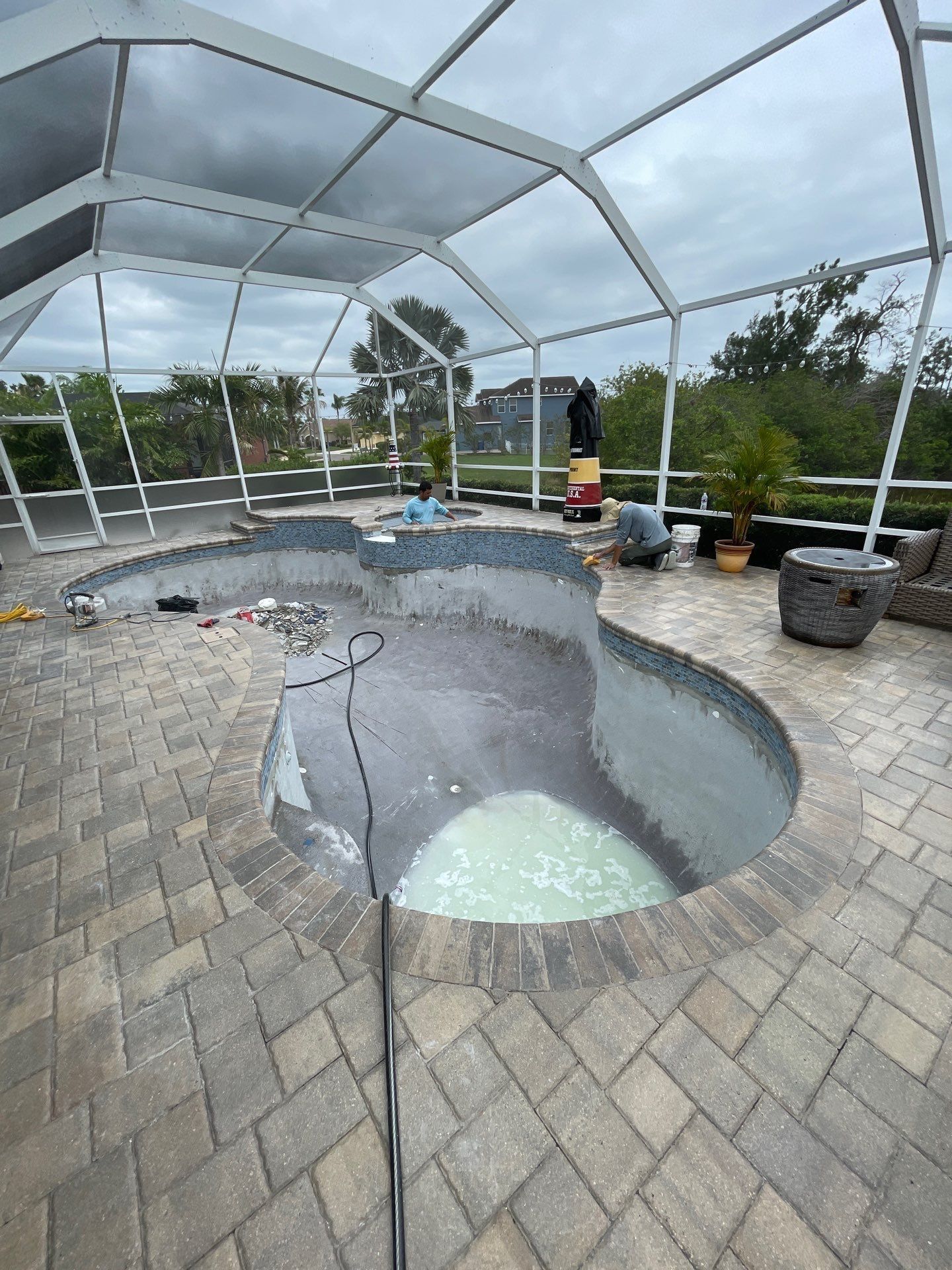 Empty screened-in pool with cracked interior and patio pavers, under a cloudy sky.