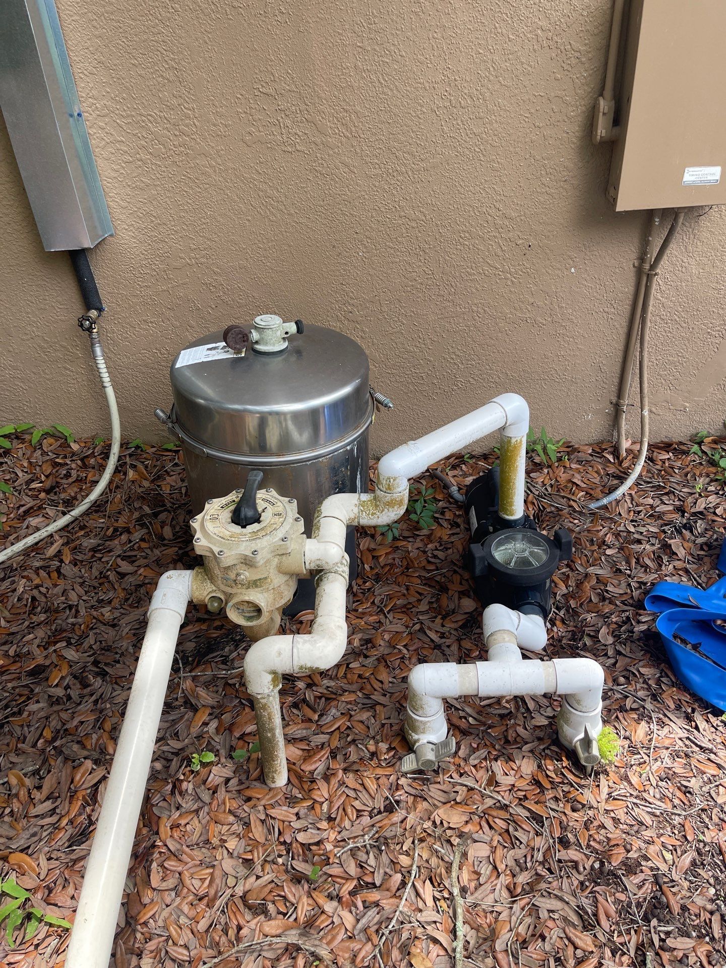 Outdoor utility pipes and valves beside a house wall, with a metal tank and mulch ground cover.