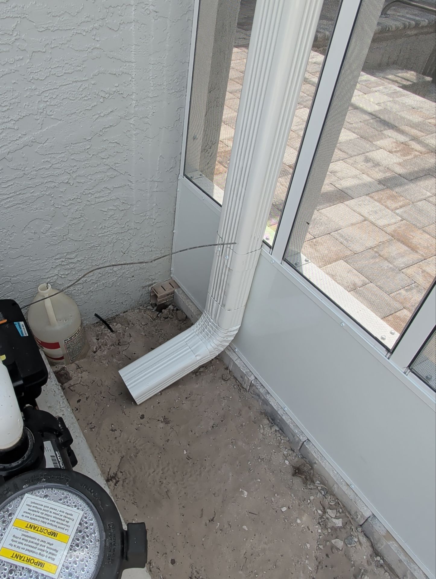 White downspout beside a glass door, ending above a concrete patio with a small gap.