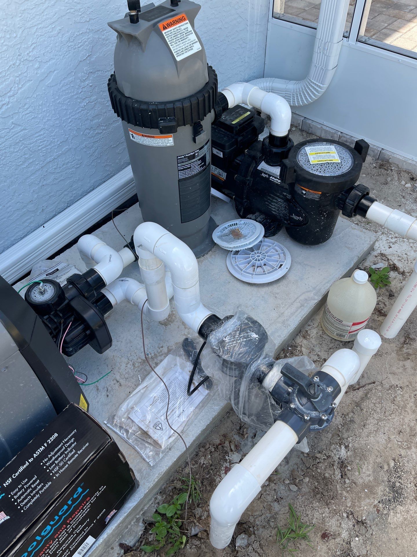 Pool pump and filter equipment with white PVC pipes and valves beside a house wall.