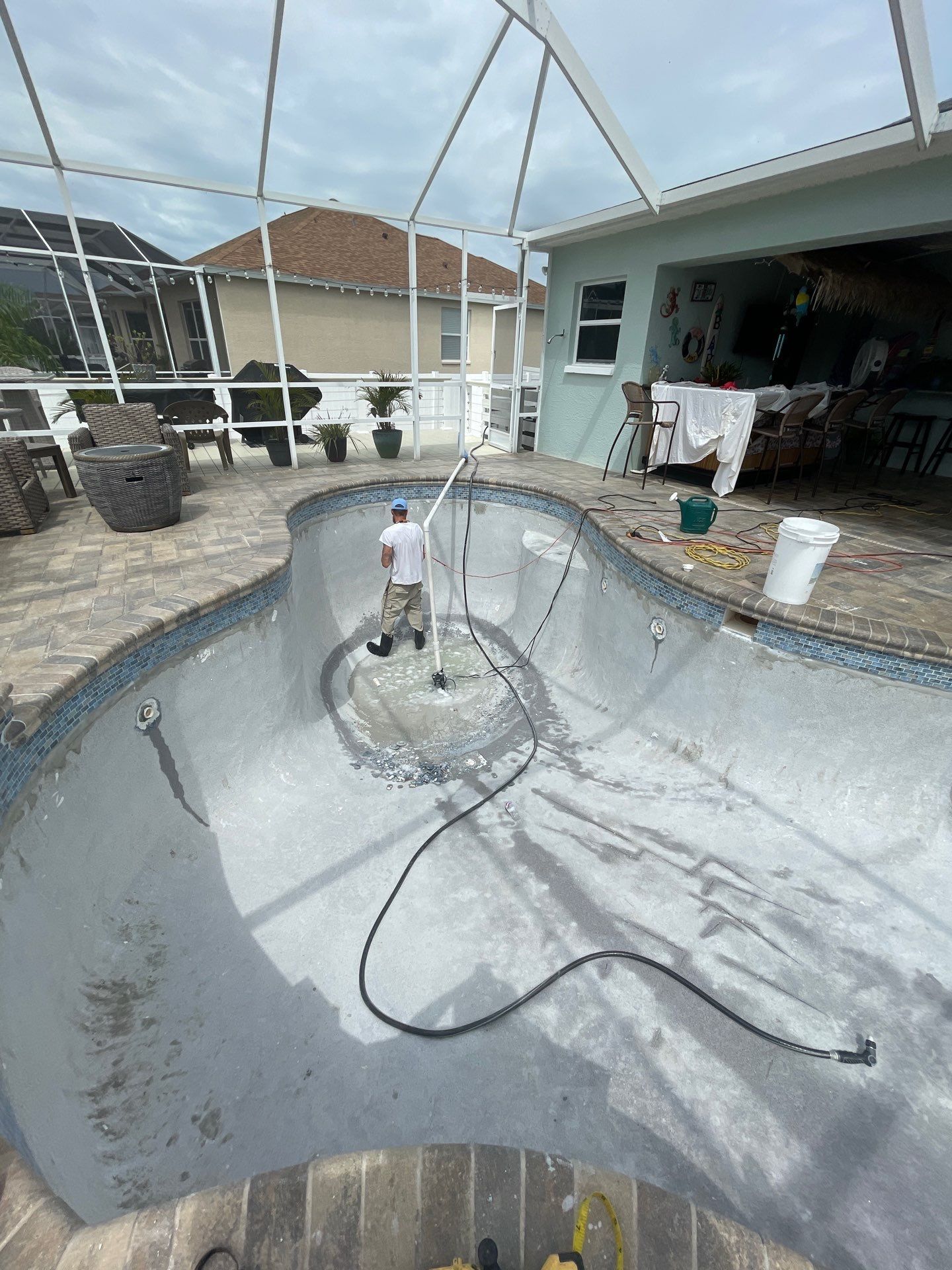 Empty in-ground pool being resurfaced, with tools and hoses inside a screened backyard patio.