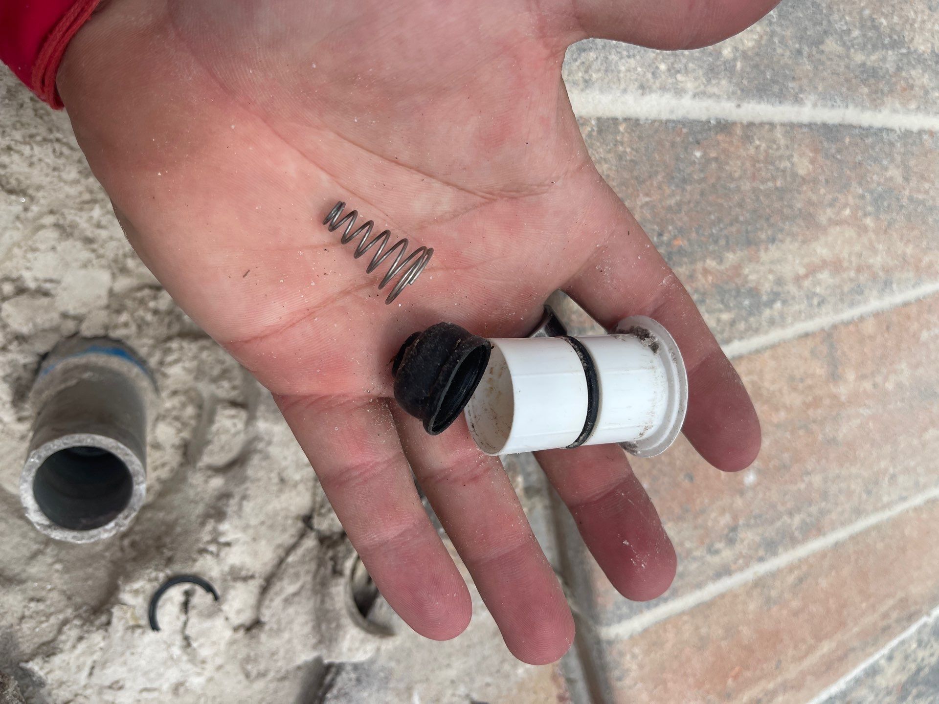 Hand holding a white plastic valve with a spring and small metal part on a tiled floor.