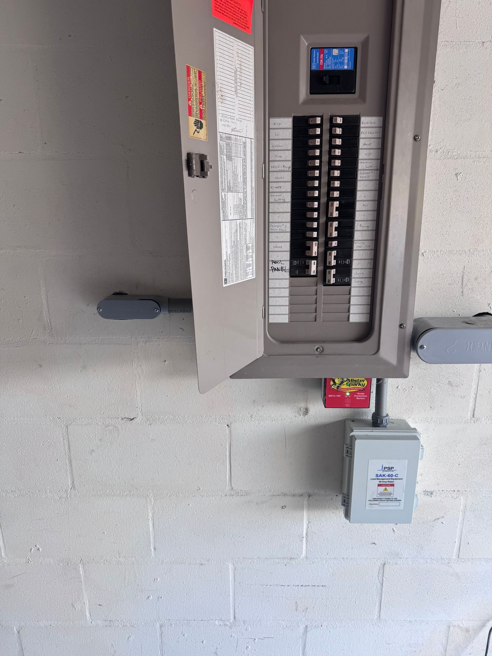 Open electrical breaker panel on a cinder block wall, with a small keypad above and a box below.
