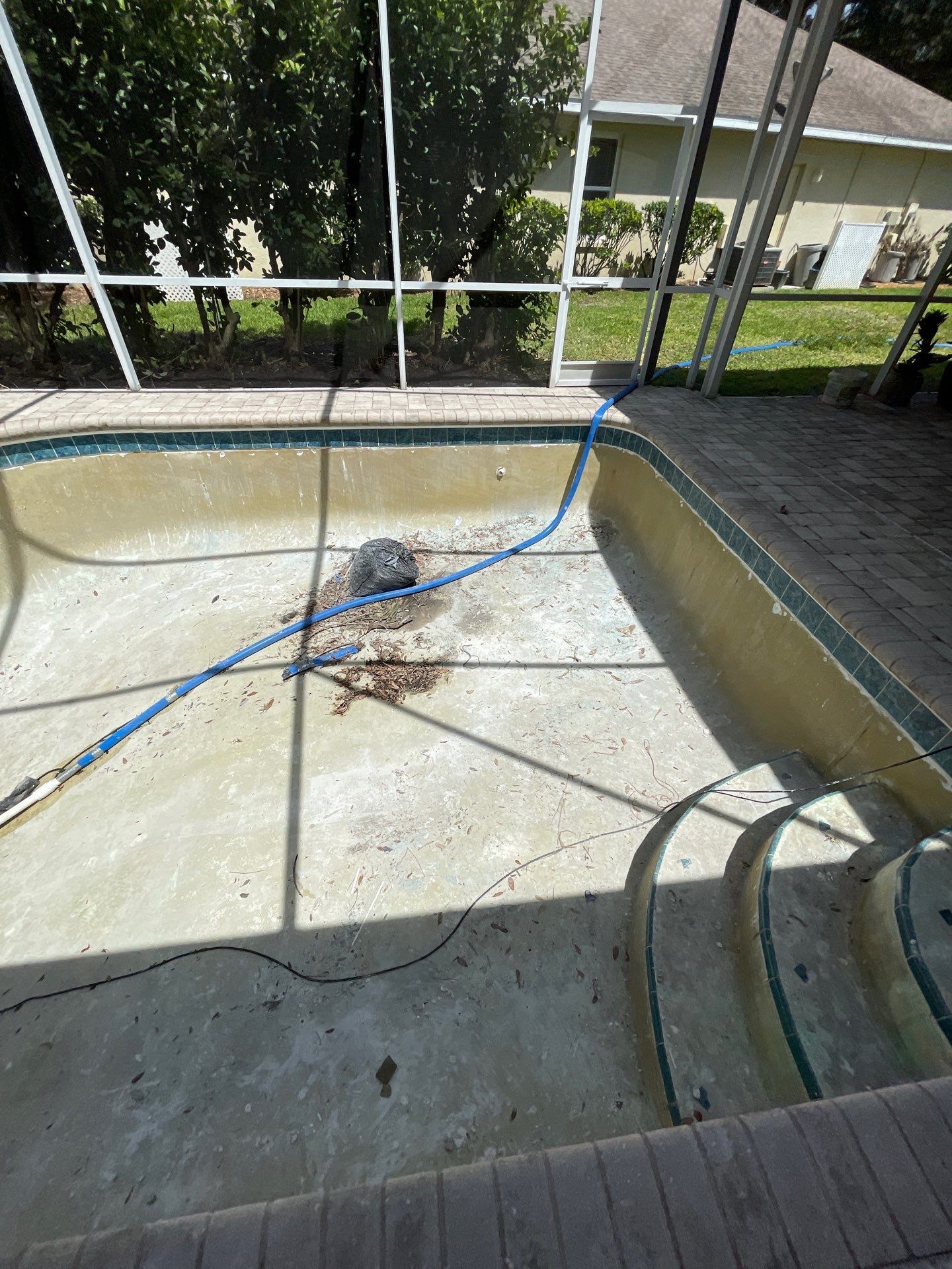 Empty drained pool with a blue hose and debris on the concrete floor near a patio.