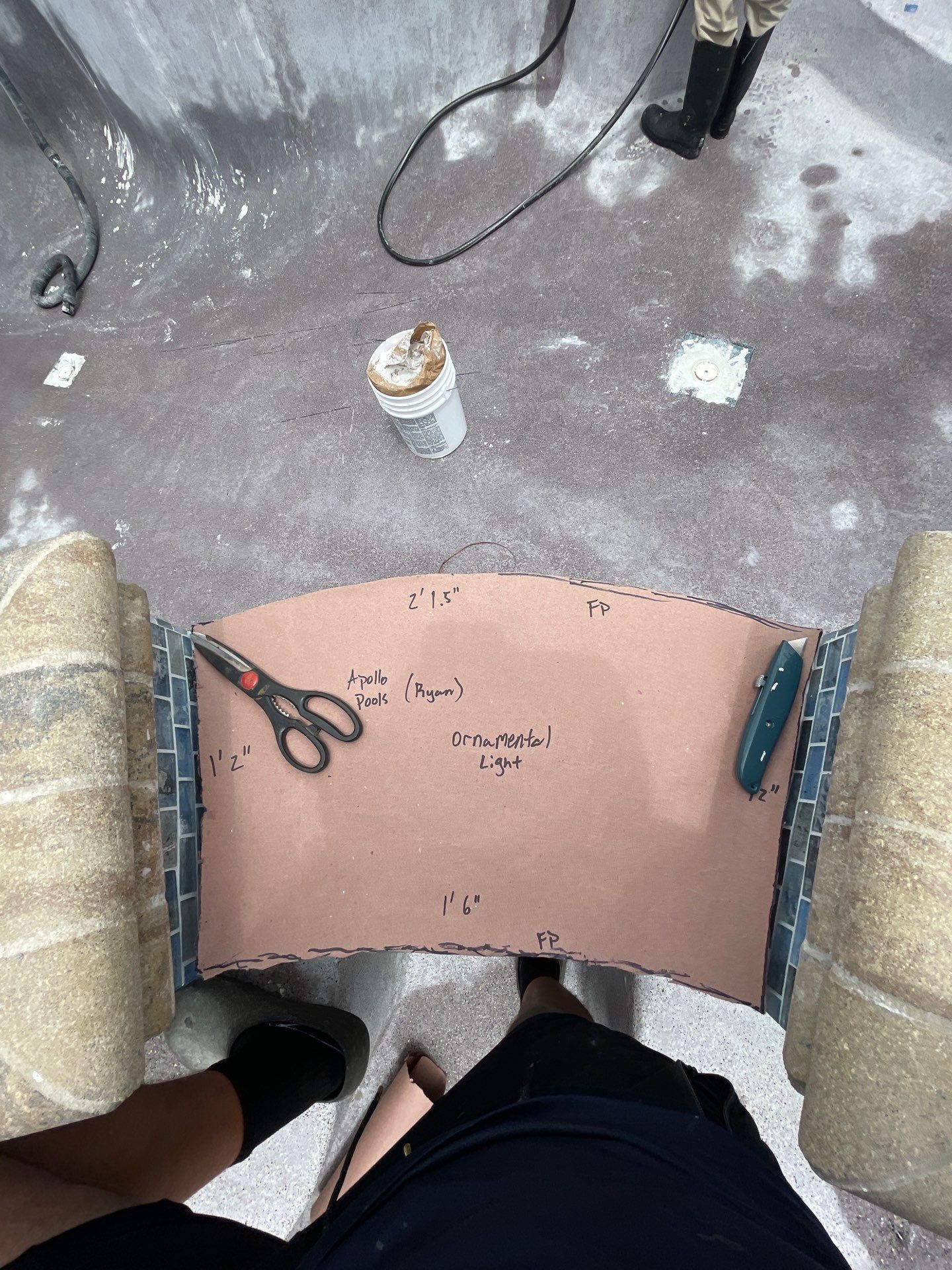 View from a rooftop: a curved pink board between concrete walls, with scissors, a bottle, and a person’s legs visible.