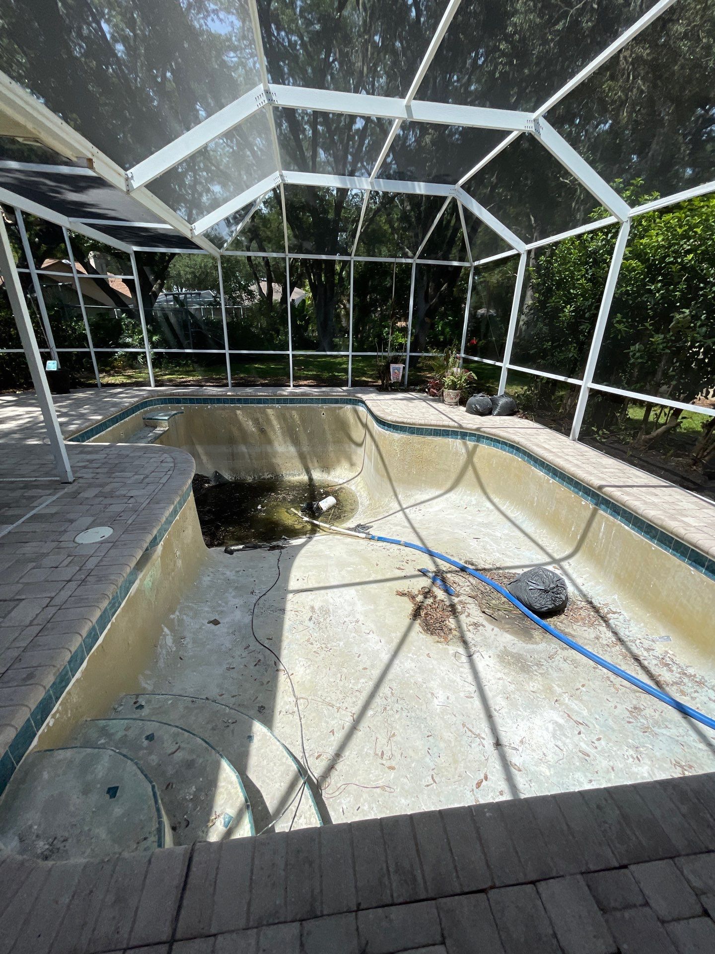 Empty screened-in pool with a drained concrete basin, debris, and a blue hose.