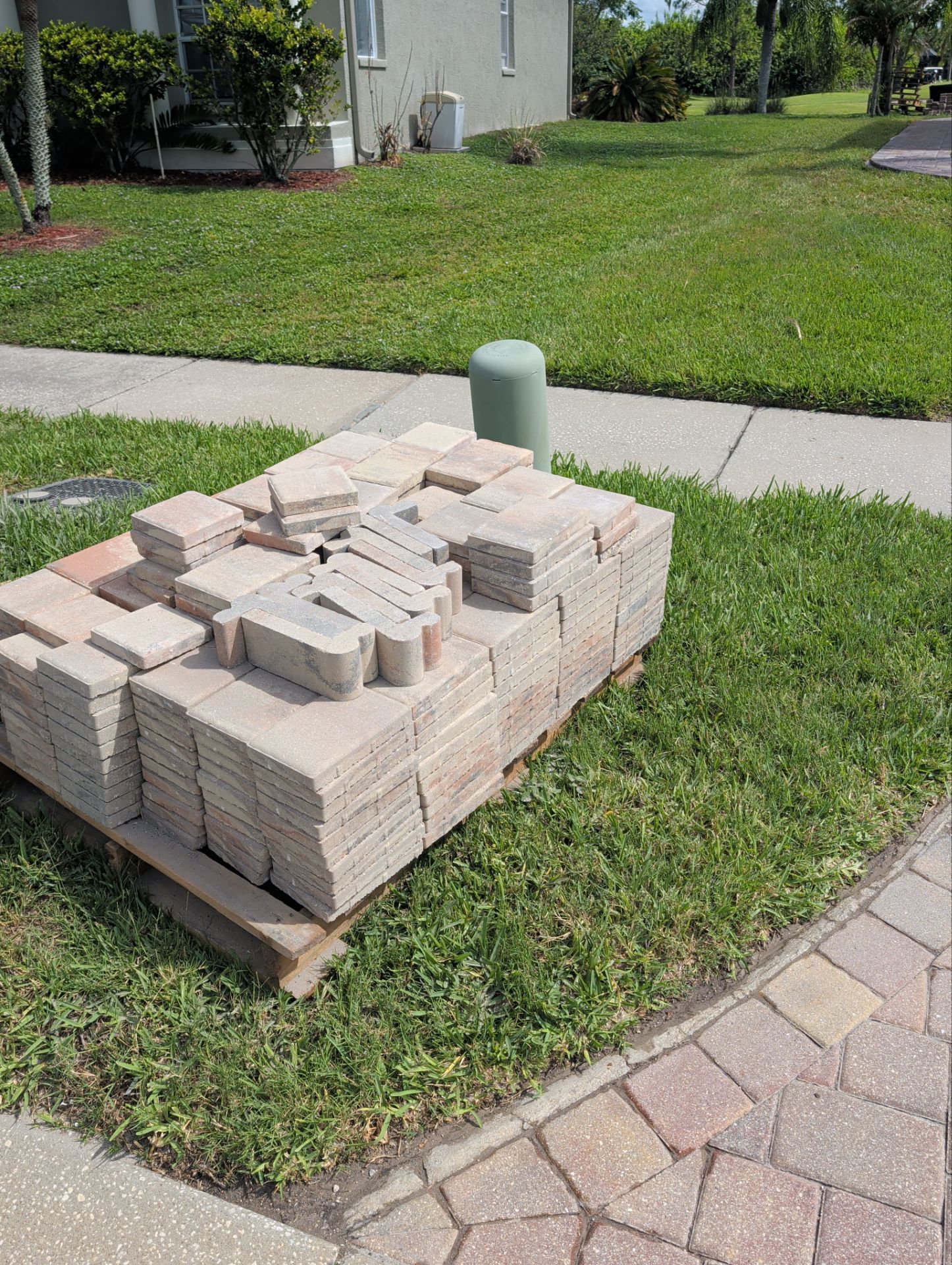 Stack of concrete pavers on a wooden pallet in a grassy yard beside a sidewalk.