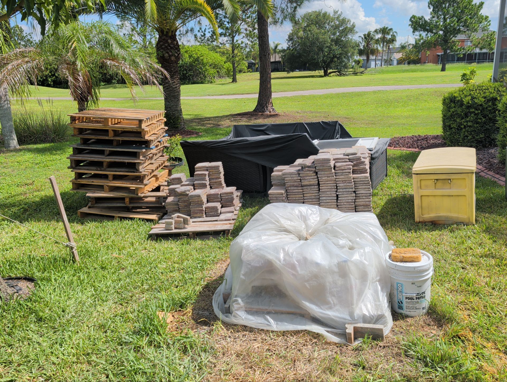 Stacks of pavers, stone blocks, and covered materials on grass in a sunny park.