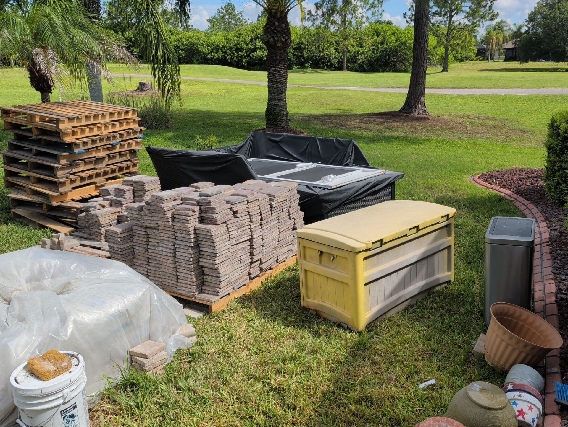 Stacked landscaping materials and debris on grass in a park-like yard, including pallets, pavers, bags, and bins.