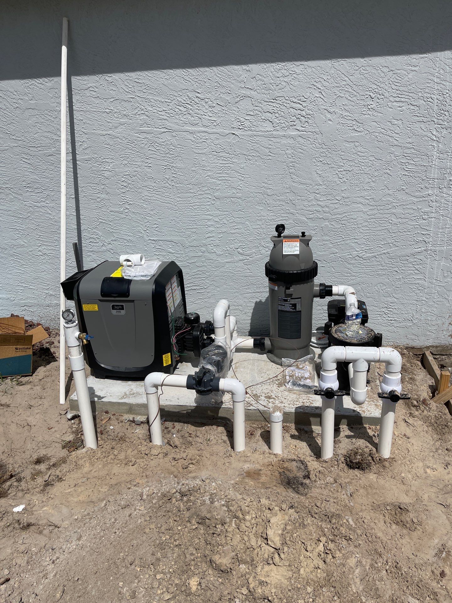 Outdoor pool equipment with pumps and piping against a gray wall, partly buried in dirt.