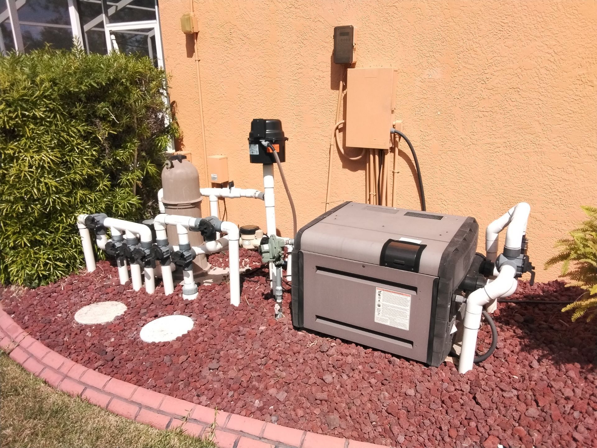 Backyard utility equipment and pipes set in red rock mulch against a tan stucco wall.
