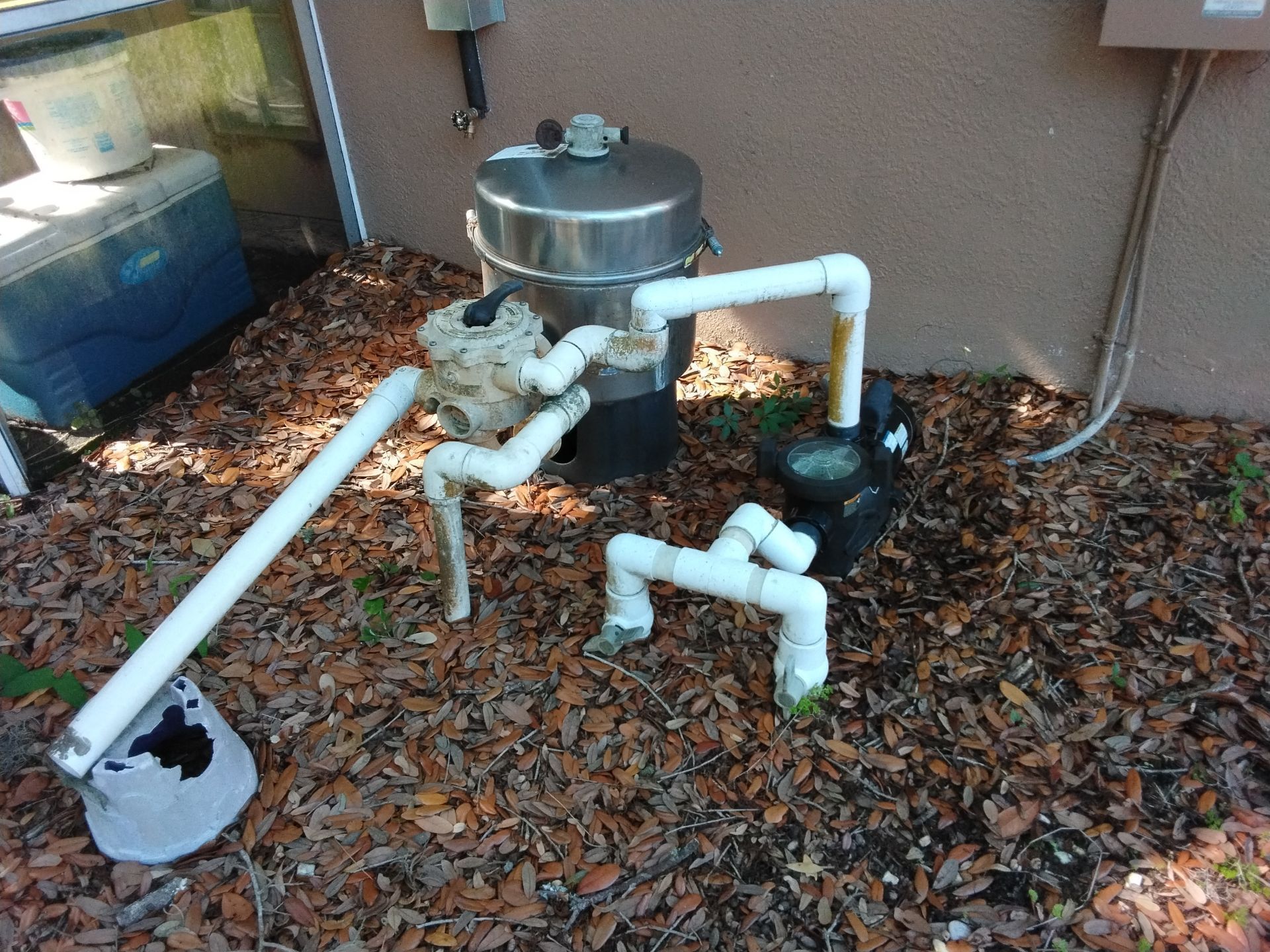Outdoor pool pump and filter system with white PVC pipes on gravel beside a wall.