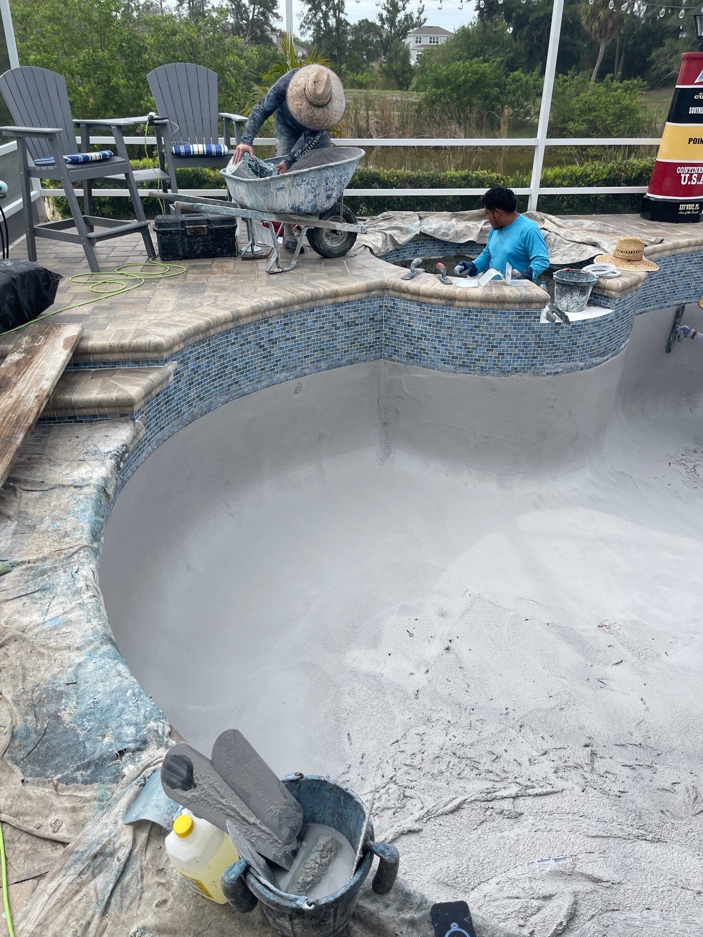 Worker painting the inside of an empty backyard pool while another person sits nearby with tools.