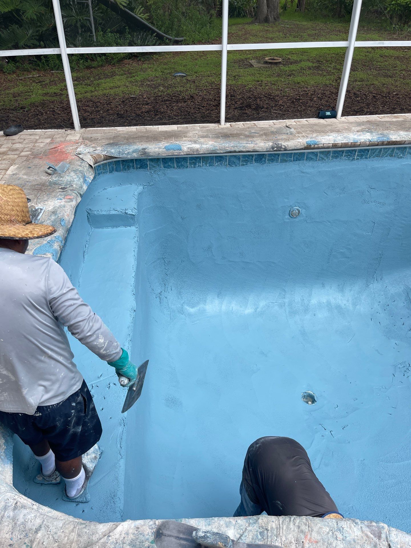 Two people painting the edge of an empty blue swimming pool outdoors.
