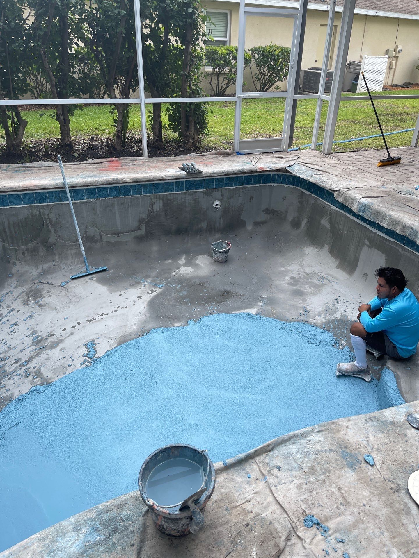 Person crouching in an empty blue swimming pool during resurfacing work with a bucket nearby.