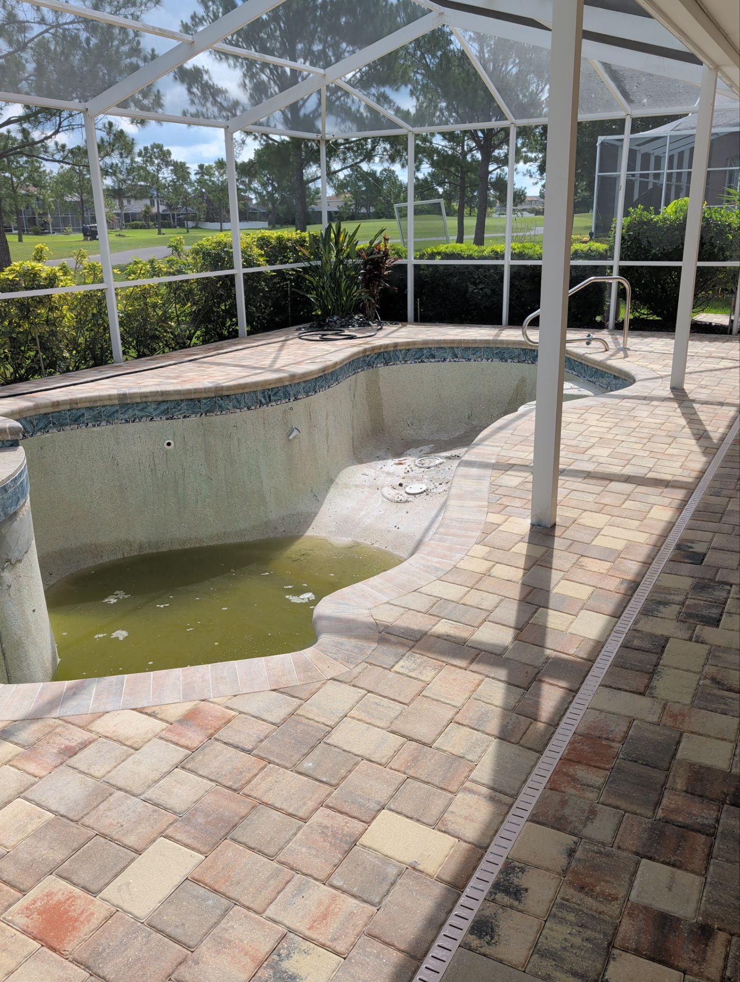 Empty screened-in pool with green water, tiled deck, and white enclosure in a sunny backyard.