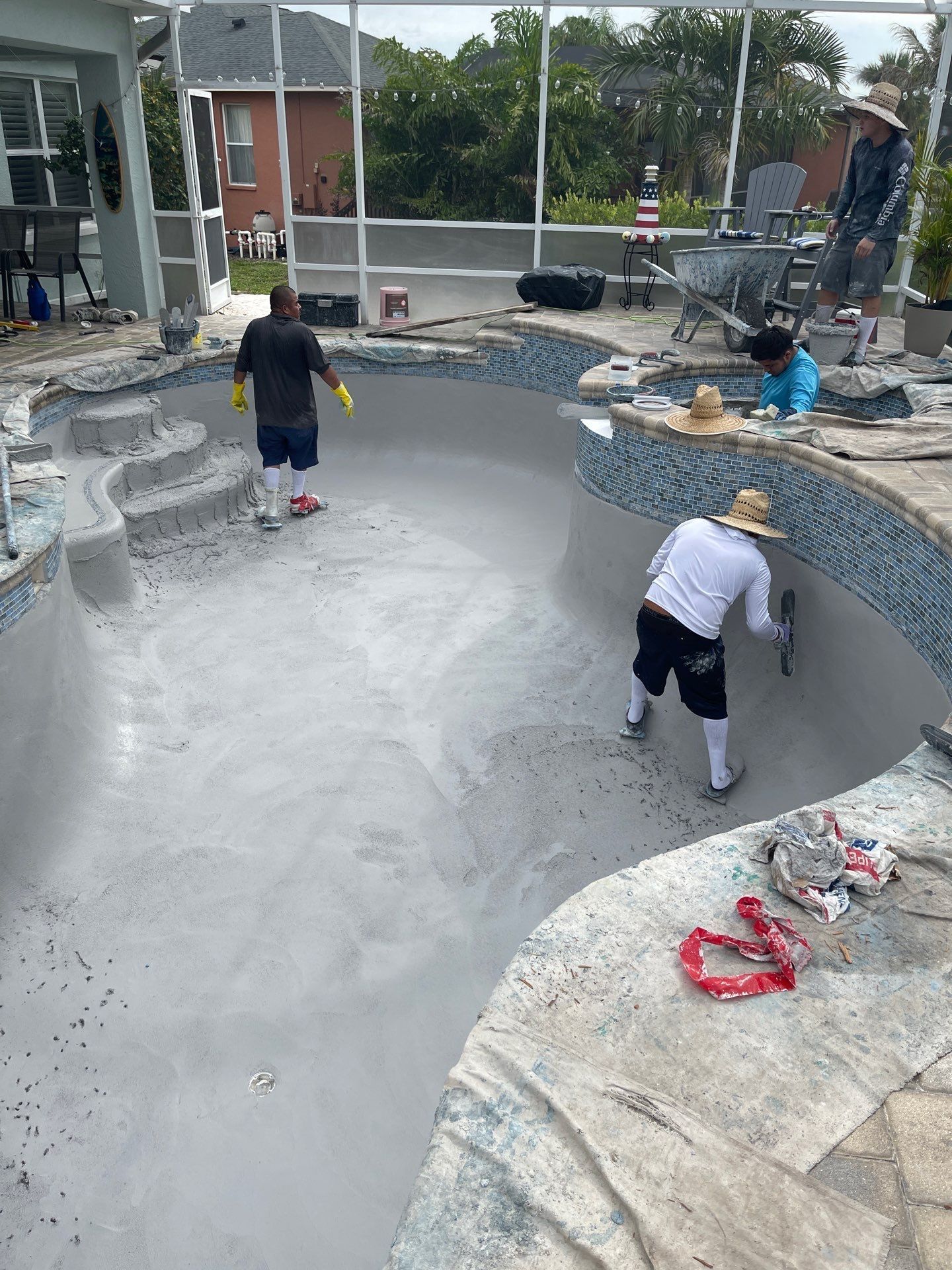 Workers resurfacing an empty backyard pool with gray cement and blue tile trim.