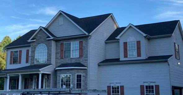 Two-story house with gray siding, stone facade, dark roof, and brown shutters against a blue sky.