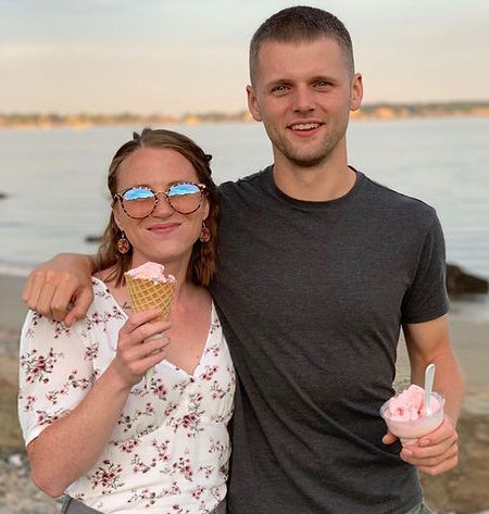 A man and a woman are standing next to each other on the beach . the woman is holding an ice cream cone.