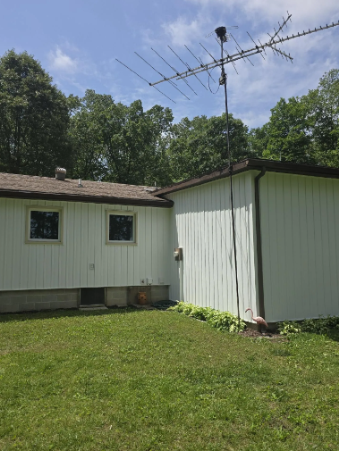 White house and shed with antenna, surrounded by grass and trees under a blue sky.
