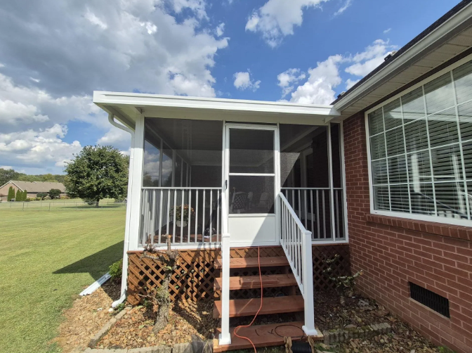 A white screen porch with stairs, attached to a brick house, under a blue sky.