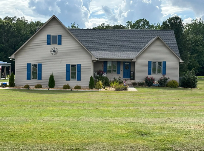 House with blue shutters, door, set in a grassy yard under a cloudy sky.
