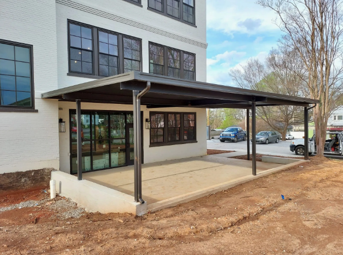 A covered outdoor patio with a concrete floor, supported by black columns, against a white brick building.