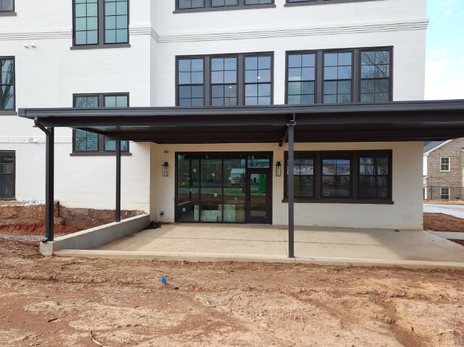 Exterior view: building with dark awning over patio. Light brick wall, black-framed windows, dirt ground.