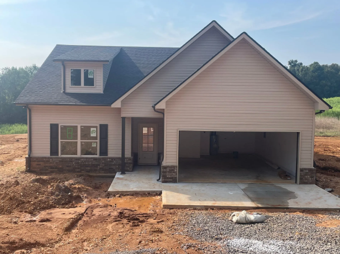 New beige house with open garage, brown roof, and construction site setting.