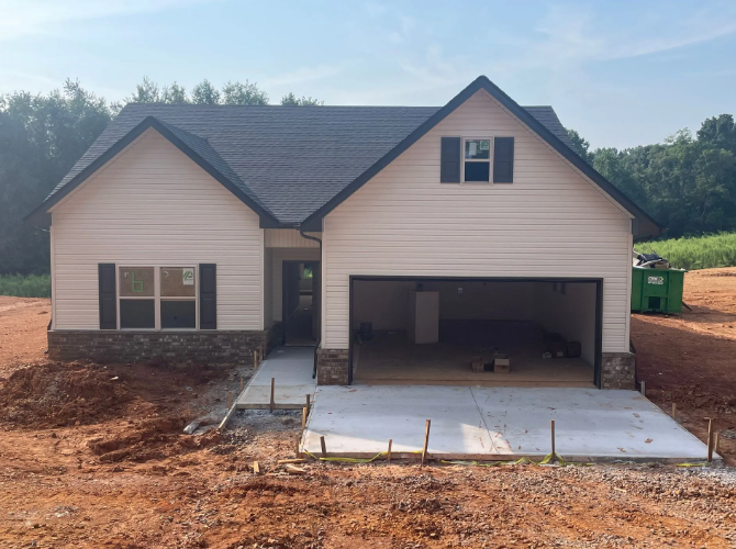 New house under construction, beige siding, dark roof, open garage, concrete driveway, surrounded by dirt.