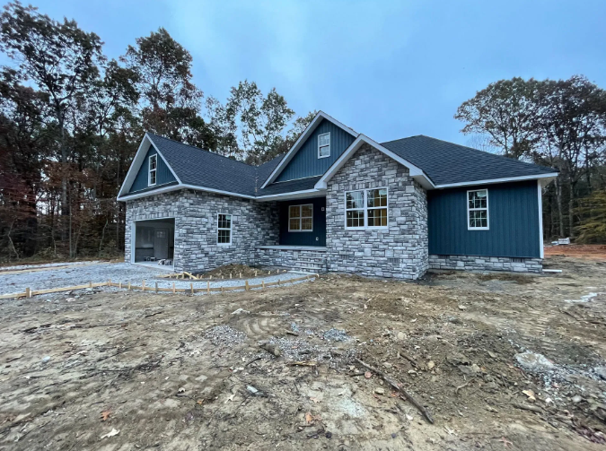 New house under construction with stone facade and blue siding, in a wooded area.