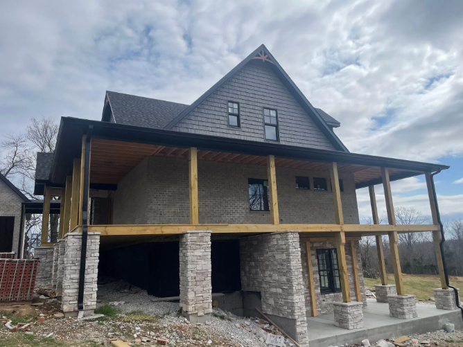 Two-story house under construction with stone columns, gray siding, and a porch.